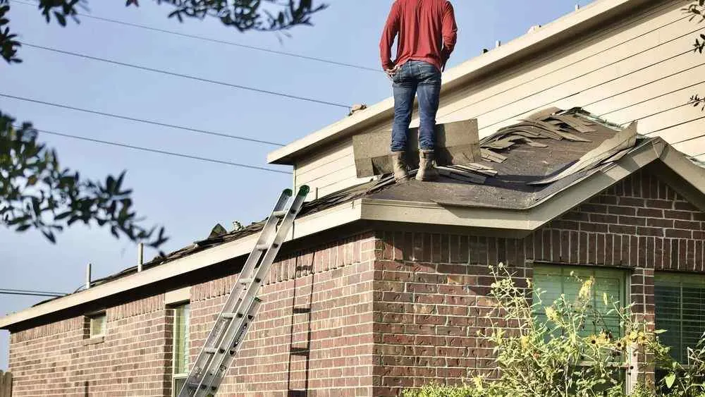 Worker standing on a damaged roof of a brick house repairing torn shingles on a sunny day.